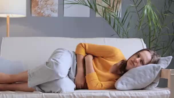 リビングルームのソファーに黄色いシャツとジーンズを着用しているUpset女性は 月経痛のクランプに苦しむ腹に手を置き 胃痛は胃を達成する慢性胃炎を持っています — ストック動画