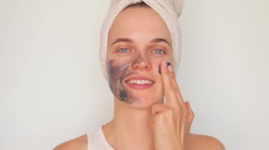 Spa yüz bakımı. Cilt bakımı. Çamur maskesi cilt sağlığı için. Güzellik ve cilt terapisi. Cilt bakımı zevki. Kafasına banyo havlusu takmış, beyaz arka planda siyah çamur maskesi takan neşeli bir kadın..