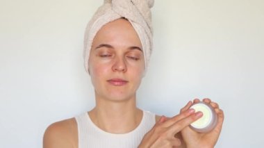 Cilt rutini. Güzel, genç bir kadın banyo havlusuna sarılmış, sabah duşundan sonra yüzüne krem sürüyor, beyaz arka planda izole edilmiş, nemlendirici, günlük cilt bakımının keyfini çıkarıyor..