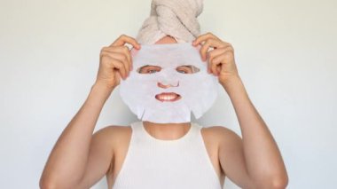Tatmin olmuş bir kadın beyaz arka planda izole edilmiş güzellik tedavisi için yüz derisine kozmetik beyaz tekstil maskesi uyguluyor..