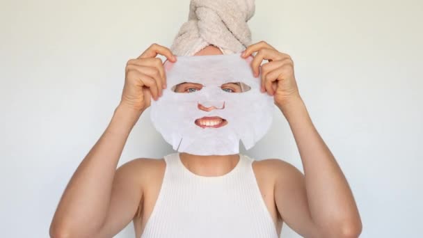 白い背景に隔離された美容処置のためのティッシュ生地の織物の織物のマスクを使用して顔の皮に化粧品の白い織物のマスクを適用する満足した女性 — ストック動画
