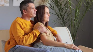 Oturma odasındaki kanepede oturan gülümseyen çift evdeki hoş sohbete dahil olmuş, mutlu genç adam ve kadın kanepede gevşeyip, romantik hafta sonları veya randevuların keyfini çıkarıyorlar..
