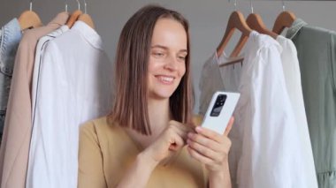 Alışverişkolik alıcı. Mobil alışveriş. E-ticaret cihazı. Banka başvurusu. Koyu renk saçlı neşeli bir kadın telefon başında moda kıyafetlerin arasında internetten yeni kıyafetler alıyor..