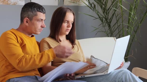 家族のビジネス用紙ワークで問題を抱えているソファー男女に一緒に座っているノートパソコンの保持文書を使用してオンラインで財政を管理する不安なカップル — ストック動画