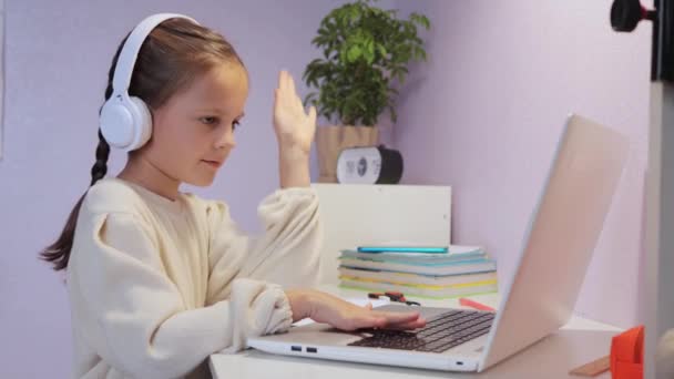 オンラインレッスン 遠隔教育について 彼女の教師のための挨拶のジェスチャーを示すフロントラップトップに机に座って白いシャツを着たピグテールを持つ少女 — ストック動画