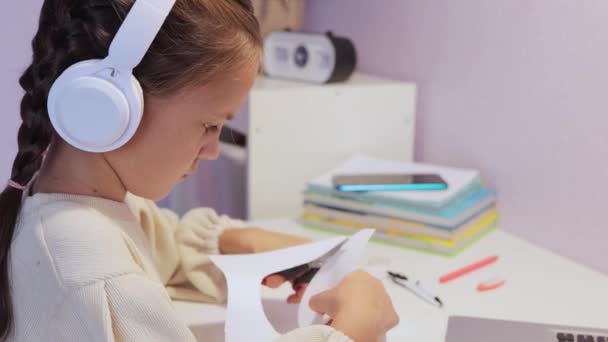 可爱的小女孩 房间里桌子上有剪刀和白纸 在家里工作 用耳机听音乐或听音频 — 图库视频影像