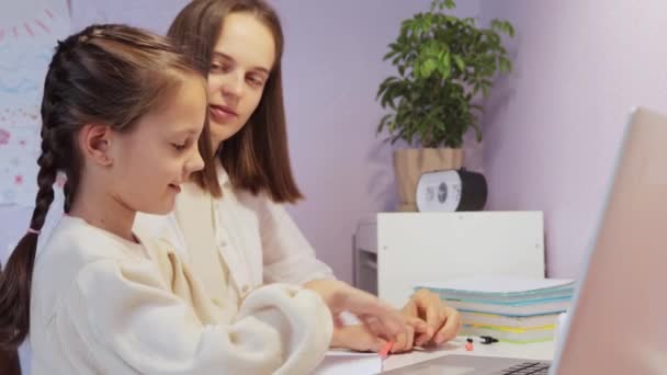 彼女の陽気な先生のチューターとラップトップの前に座っているピグテールと笑う小さな少女は 勤勉な学生の母親が娘の仕事のやり方を説明することを賞賛します — ストック動画