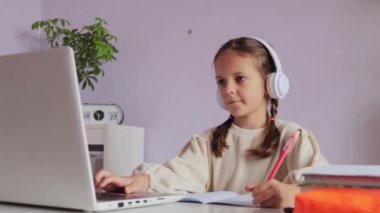Saçları örgülü, beyaz tişörtlü, kulaklıklı, kulaklıklı ev ödevi yapan bir kız..