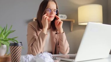 İnternet işi. Bir bilgisayar kullanarak. Resmi stil ceket giyen, yorgun, kahverengi saçlı bir kadın laptopta uzun saatler çalışıyor baş ağrısı çekiyor modern ışık ofisinde acı veren şakaklara masaj yapıyor..