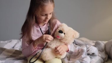 Şirin kahverengi saçlı küçük kız evde yatağında oturmuş oyuncak ayısını muayene etmek için stetoskop kullanarak doktorculuk oynuyor..