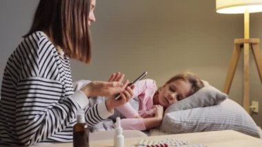 Üzgün hasta bir kız yatağında dinlenirken annesi doktorla konuşuyor. İnternette haplar ve burun spreyi gösteriyor. Modern akıllı telefondaki video görüşmesi sırasında doktorla internet danışmanlığı yapıyor..