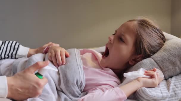 医学的な病気 子供の健康について 喉の痛みに苦しむ小さな少女は 彼女の病気の娘を世話し サポートしているベッドに横たわるトニルスプレーに広く口を開きます — ストック動画