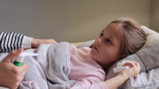 国内治療について 小児病について 子供の熱がする 病気の少女は 母親が彼女を助けながら トニルスプレー開口口でそれを治療する喉の症状を持つベッドに横たわる熱を持っています — ストック動画