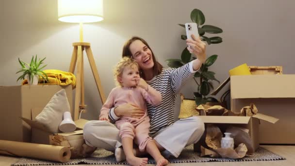 ホーム移転の幸福について 笑顔のコーカサス人の母親と赤ちゃんの娘は 移動日にセルフィーやビデオ通話をするものとダンボール箱で新しいアパートを移転しました — ストック動画