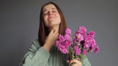 Polen mevsimi. Rhinitis reaksiyonu. Kasımpatı alerjisi. Mevsimsel hastalıklar. Hasta kadın, yeşil örülü gömlek giyip elinde buket tutuyordu. Gri arka planda soyutlanmış, boynu kaşınıyordu.