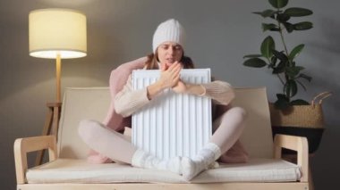 Mevsimsel sıcaklık değişir. Kışın ısınıyorum. Şapka ve palto giymiş üzgün bir kadın kanepede oturuyor ve ısınmak için avuçlarını ovuşturup ısıtıcıyı kucaklıyor..