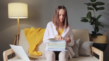 Yetişkinler evde ders çalışıyor. Öğrenci teste hazırlanıyor. Uzaktan eğitim dersleri. Oturma odasında, bilgisayar ve kitaplarla kanepede oturmuş cevap arayan genç bir kadın..