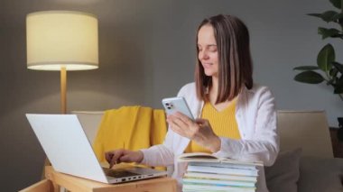 Akıllı telefon kullanan gülümseyen genç bir kadın akıllı telefondan bilgi toplarken dizüstü bilgisayarda klavye hazırlarken ya da üniversite ya da test denemesi için kompozisyon hazırlarken.