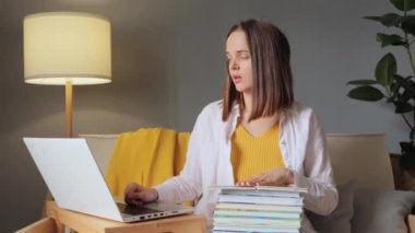 Laptoptan öğreniyorum. Çevrimiçi üniversite dersleri. Evde ders çalışan yorgun genç bir kadın oturma odasında bilgisayar ve kitaplarla oturup rahatsız edici bir poz vererek boyun ağrısı çekiyor..