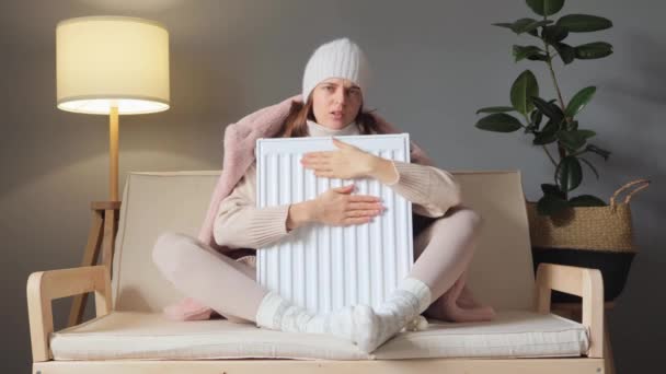 ホームヒーティングシーズン 冬のインテリア暖かさ エネルギー効率の良い暖房 ソファーに座ってヒーターラジエーターを受け入れるキャップとコートを身に着けている悲しい女性 — ストック動画