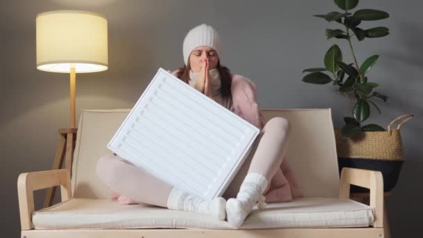 家を暖めること 経済的な暖房の解決 バッテリー駆動の暖かさ 冷たい悲しい女性は ソファーに座ってヒーターラジエーターを受け入れる帽子とコートを着用しています — ストック動画