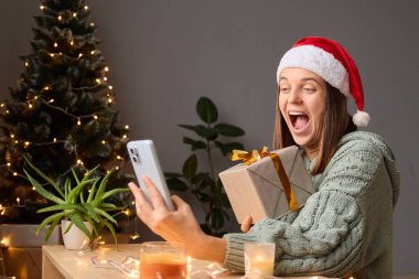 Aşırı neşeli, örgü gömlekli ve Noel Baba şapkalı bir kadın elinde Noel Baba kutlamasıyla evde oturmuş Noel 'i internetten kutluyor ve neşeli bir şekilde bağırıyor..