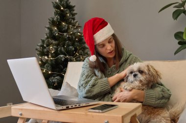 Çekici bir kadın Noel 'i köpeğiyle birlikte dizüstü bilgisayarında film izleyerek kanepeye oturup Noel Baba şapkası takarak geçiriyor..