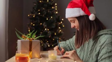 Noel Baba şapkası takan ve Noel dilekleri yazan bir kadın yeni yıl bayramı için hediye listesi hazırlıyor..