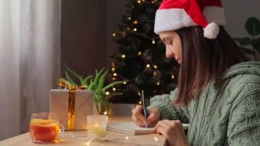 Gelecek için motivasyon. Genç yetişkin bir kadın örgü gömlek ve Noel Baba şapkası takıyor. Yılbaşı için notlar yazıyor. Evdeki defterde, Noel ağacı planlaması yapıyor..