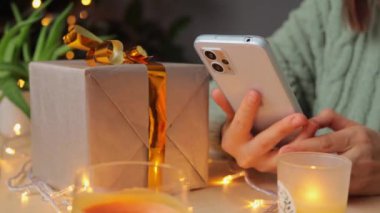 Örgü gömlekli tanınmayan bir kadın arka planda Noel ağacıyla süslenmiş bir masada oturuyor cep telefonuyla internetten alışveriş yapıyor..