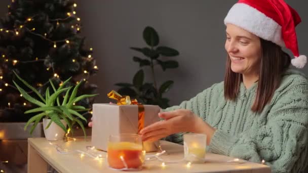 身穿针织衬衫 头戴圣诞帽 面带微笑的女人坐在桌旁 手里拿着包装好的礼品盒 喜形于色 欢欢喜喜地庆祝寒假 — 图库视频影像