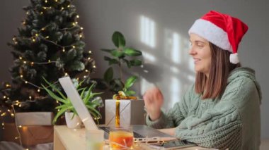 Dost canlısı, yeşil örgü gömlekli ve Noel Baba şapkalı bir kadın Noel süslemeli odada el sallayarak el sallıyor, gülümsüyor ve mutlu bir şekilde Noel 'i kutluyor..