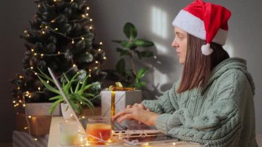 Noel Baba şapkası takan ve örgü ören güzel bir kadın süslü bir odada oturmuş dizüstü bilgisayarda klavyesini daktilo ediyor ve yılbaşı gecesi online iş için akıllı telefonu kullanıyor..