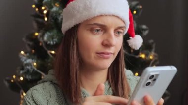 Noel Baba şapkası takan şaşırmış bir kadın dekoratif iç kayışlarda Noel ağacının yakınındaki telefonu kullanarak rahatsız edici ve şok edici bir şey görüyor..