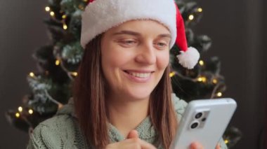 Mutlu, güler yüzlü, Noel Baba şapkası takan neşeli bir kadın dekoratif iç mekanda Noel ağacının yanındaki telefonu kullanarak Noel 'de mutlu duygularla cep telefonu kullanıyor..