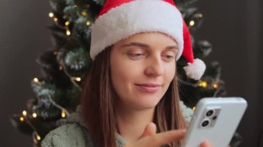 Noel Baba şapkası takan neşeli bir kadın dekoratif iç mekanda Noel ağacının yanındaki telefonu kullanarak uzun zamandır beklenen sevindirici haberleri kutluyor..