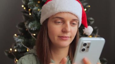 Noel Baba şapkası takmaktan yorgun düşmüş bir kadın. Dekoratif iç mekanda Noel ağacının yanında telefon kullanıyor. Sosyal ağları kontrol ediyor, esnemekten yoruluyor..