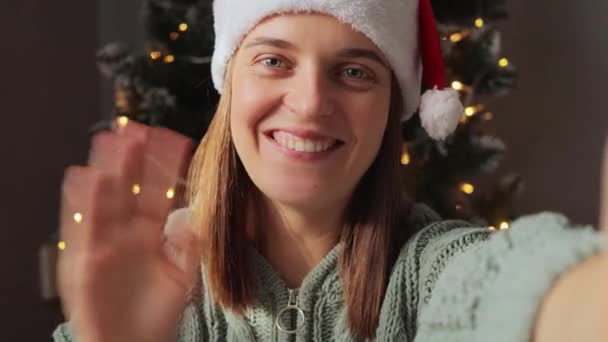 笑容满面 头戴圣诞帽的女人挥手打招呼 微笑着对着摄像机记录下的圣诞祝贺视频 — 图库视频影像