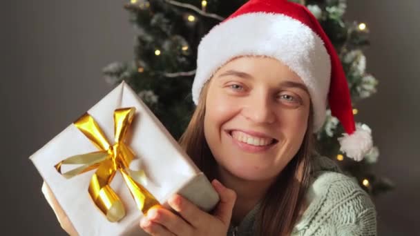 笑容满面的年轻女子戴着圣诞礼帽 在装饰过的房间里摆出礼物盒 背景上挂着圣诞树 表达着幸福和喜庆的心情 — 图库视频影像