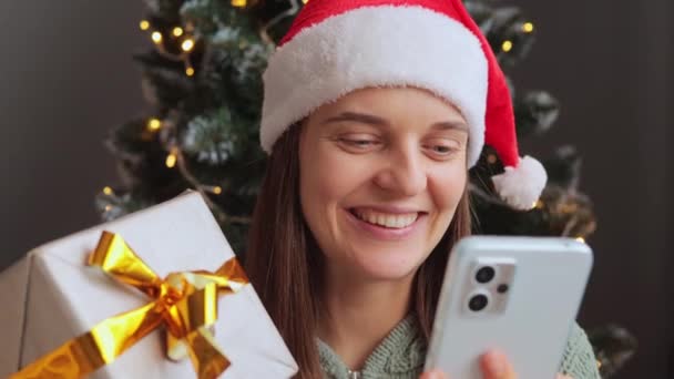 快乐的女人带着快乐的笑容 在智能手机上放着视频 看着摆放着快乐表情的小玩艺儿 恭贺新年给她看圣诞礼物 — 图库视频影像