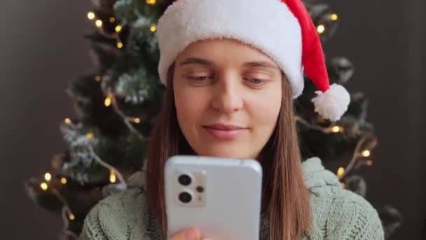 サンタクロースの帽子をかぶっている驚くべき女性は ネガティブな感情を表現するソーシャルネットワークで悪いニュースを読む装飾的な内装の木の近くで電話を使用して驚きました — ストック動画