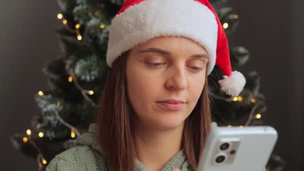 戴着圣诞帽的打哈欠的女人在圣诞树边用手机在装饰性室内感觉疲劳 因为网上打滚时感到困倦 — 图库视频影像