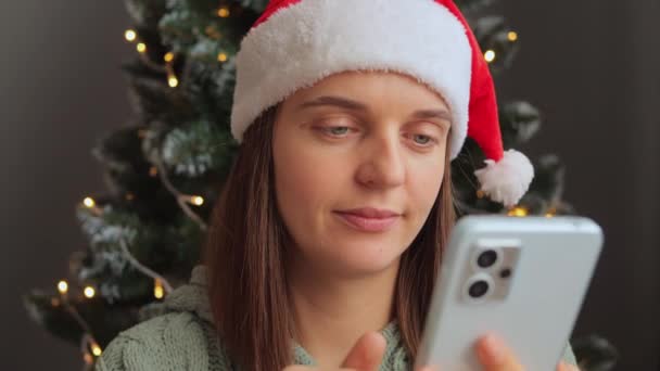 疲惫困倦的女人头戴圣诞帽 在装饰性室内的圣诞树旁打电话 检查社交网络 打呵欠累坏了 — 图库视频影像