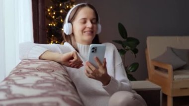 Genç, kahverengi saçlı bir kadın Noel ağacının yanında oturmuş akıllı telefon takmış, kulaklık takmış neşeli müzik dinliyor ve rahatça dans ediyor..