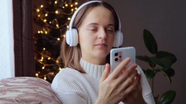 Sakin, rahat, kulaklık takan, müzik dinleyen bir kadın. Noel atmosferinde mutlu ve mutlu. Yeni yıl ağacının yanındaki koltukta oturuyor..