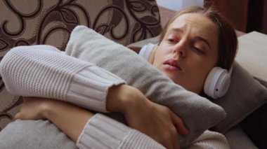Oturma odasında müzik dinleyen ve yastığı kucaklayan, endişeli yüzlü, kulaklıklı beyaz bir kadın vardı..