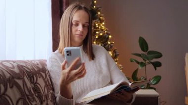 Gülümseyen neşeli kadın, kış tatillerinde Noel ağacının yanında oturmuş cep telefonuyla mesaj okuyor ve kitap okuyor ya da üniversite derslerine hazırlanıyor..