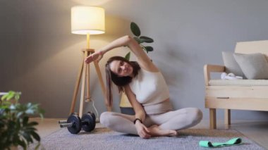Sağlıklı olmak için evde çalışıyorum. Sağlıklı yaşam tarzı ve zindelik. Alıştırma yapmadan önce evde yoga yapan çekici genç bir kadın kollarını ve yanlarını esneterek sağlığını ve sağlığını geliştiriyor..