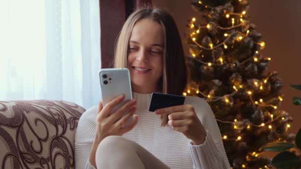 クリスマスショッピング 新年のお祝い Masツリーの近くのソファーに座っているギフトの支払いのための携帯電話のクレジットカードからデータを入力する陽気な茶色の髪の女性 — ストック動画