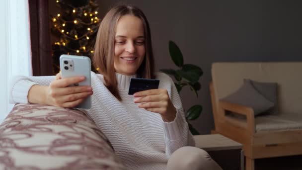 Masツリー近くのソファーに座って クリスマスプレゼントを買ってクレジットカードから支払い情報を入力するポジティブな笑顔の茶色の髪の女性 — ストック動画